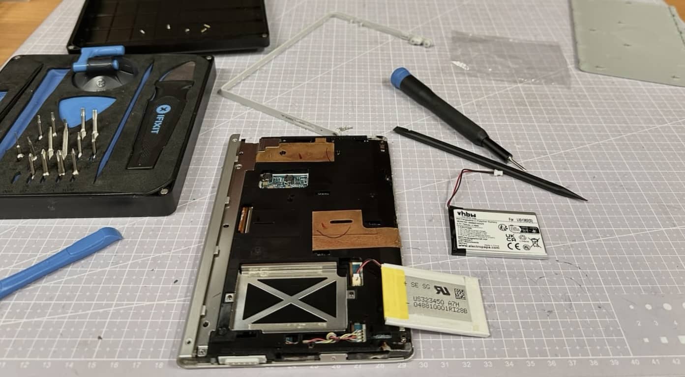 The back of the PRS-300 is open and shows the old battery. The new aftermarket battery from vhbw lies next to it. A small ifixit set and a few tools, as well as parts of the eReader are strewn around.